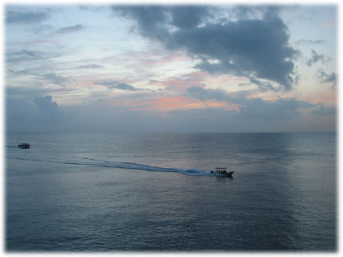 Two boats move across water with sunset behind them